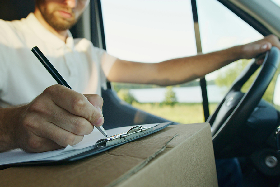 Man sitting in a vehicle with one hand on the wheel and the other filling out paperwork on top of a cardboard box.